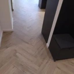 Ułożona podłoga z paneli w jodełkę francuską, widoczny fragment czarnej zabudowy meblowej z białą listwą oraz fragment białej ściany z listwą przypodłogową.