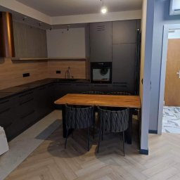 HomeStep - Drzwi Panele Poznań - Nowoczesna kuchnia z ciemnymi szafkami, drewnianym blatem i podłogą ułożoną w jodełkę, widok z perspektywy podłogi, z fragmentem nieukończonej posadzki po lewej stronie.