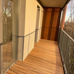 HomeStep - Drzwi Panele Poznań - Balkon z drewnianą podłogą w ciepłym odcieniu, metalowa balustrada, widok na drzewa w tle, popołudniowe słońce rzuca cienie.