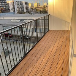 HomeStep - Drzwi Panele Poznań - Nowoczesny balkon z deskami kompozytowymi w odcieniach brązu, metalowa balustrada, widok na budynki i żurawie budowlane w tle, ciepłe światło słoneczne.
