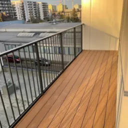 Nowoczesny balkon z deskami kompozytowymi w odcieniach brązu, metalowa balustrada, widok na budynki i żurawie budowlane w tle, ciepłe światło słoneczne.