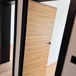 HomeStep - Drzwi Panele Poznań - Otwarta, fornirowana skrzydło drzwiowe w czarnej ościeżnicy, widok na sąsiednie pomieszczenie z drugimi drzwiami w czarnej ramie, drewniana podłoga.