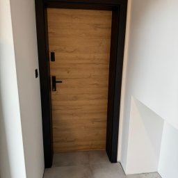 HomeStep - Drzwi Panele Poznań - Drewnopodobne skrzydło drzwiowe z czarną ościeżnicą i klamką, widoczne od strony wewnętrznej, z kluczem w zamku.