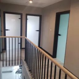 Korytarz z trzema drzwiami o czarnych framugach: dwa białe, jeden turkusowy; balustrada z drewnianą poręczą i czarnymi, pionowymi szczebelkami; na dole widoczna piła ukośnica na stole.