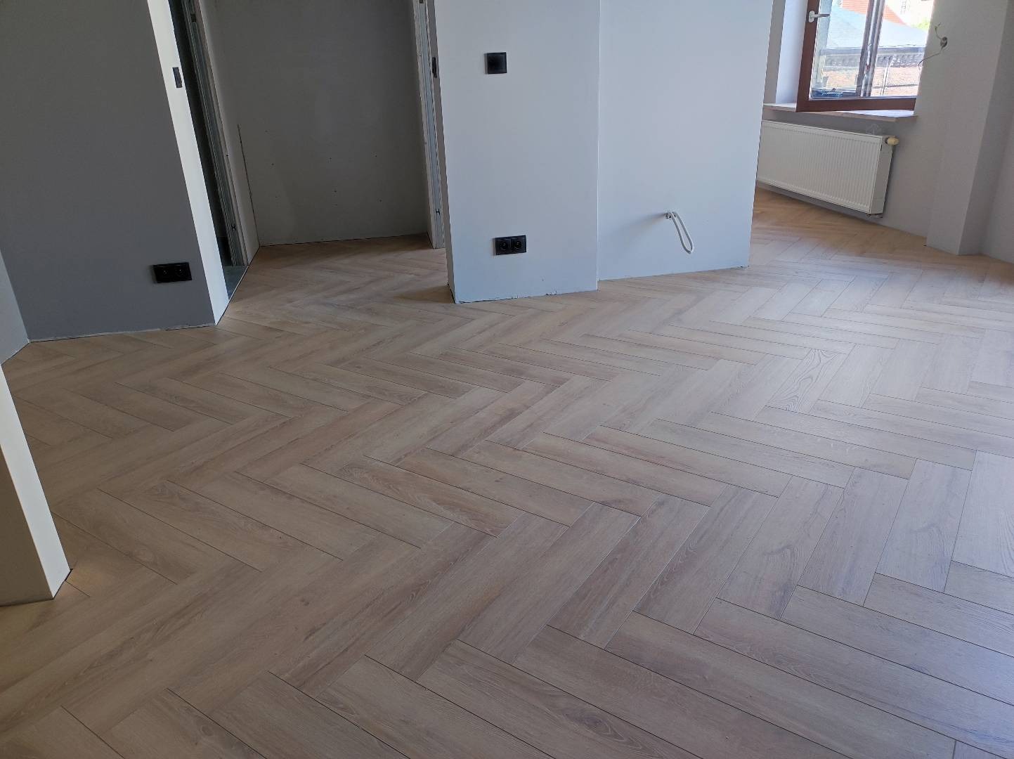 Ułożona podłoga z paneli w jodełkę w jasnym odcieniu drewna, widoczne szare ściany w surowym stanie, czarne gniazdka elektryczne i grzejnik pod oknem.