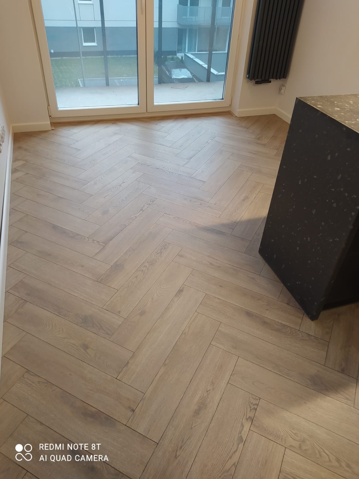 Ułożona podłoga z paneli w jodełkę francuską, widok na okno balkonowe i grzejnik.