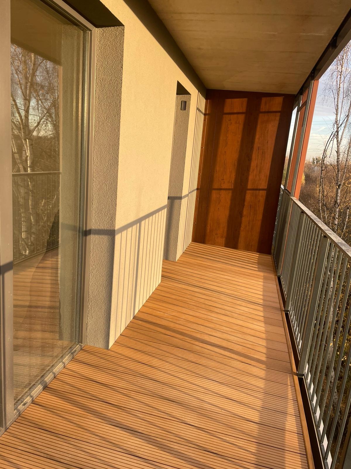 Balkon z drewnianą podłogą w ciepłym odcieniu, metalowa balustrada, widok na drzewa w tle, popołudniowe słońce rzuca cienie.