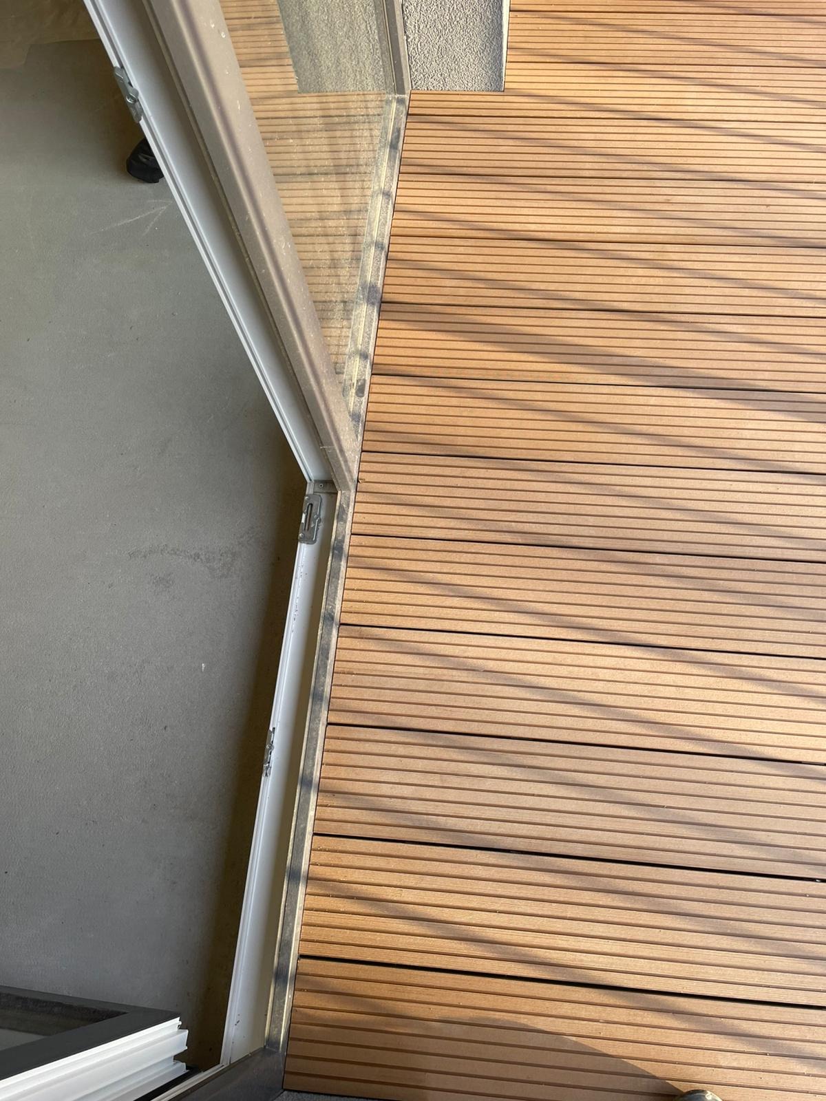 Fragment podłogi z desek kompozytowych na tarasie, widok z góry, z widocznym progiem drzwi balkonowych i fragmentem betonowej posadzki.