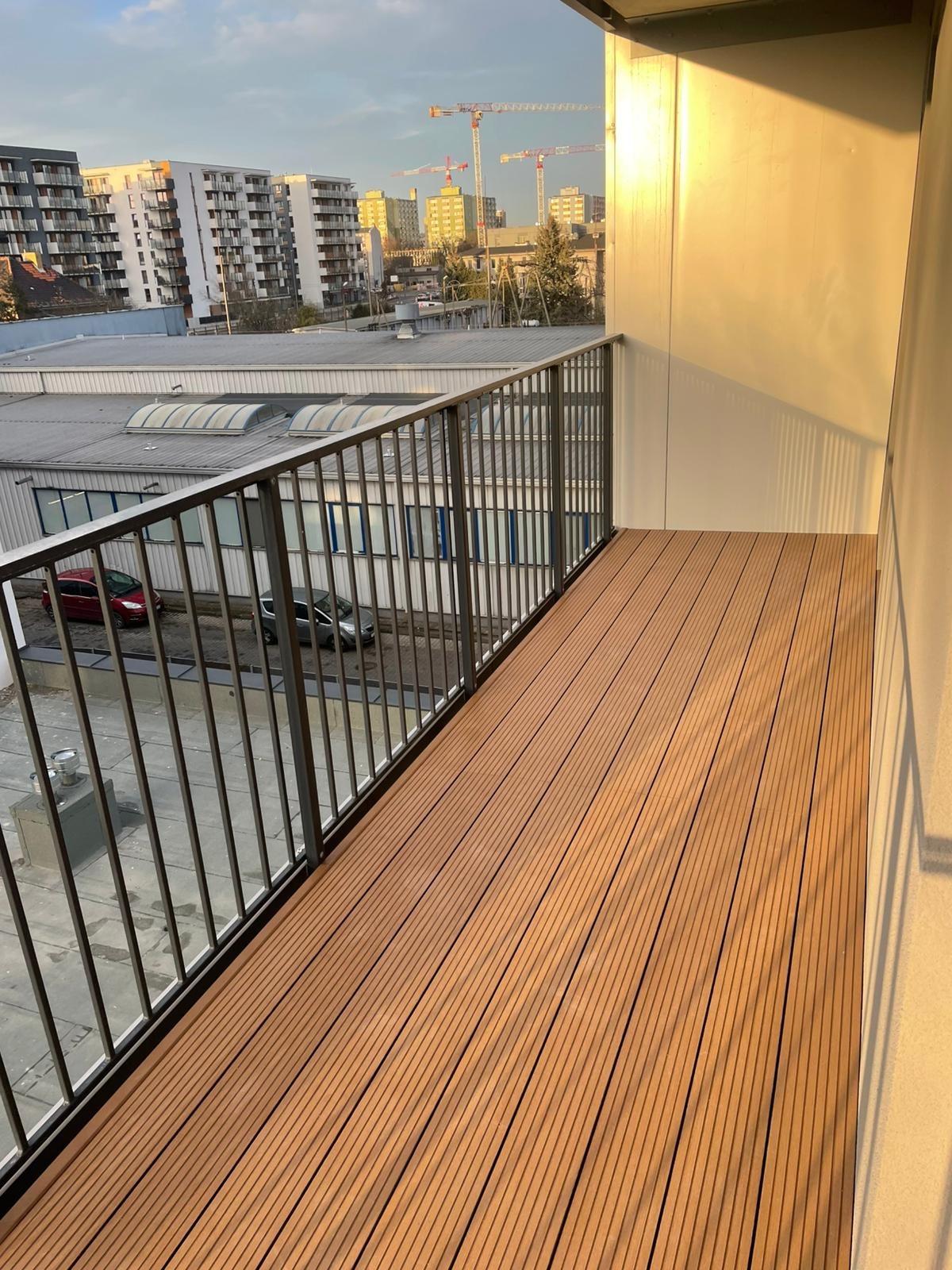 Nowoczesny balkon z deskami kompozytowymi w odcieniach brązu, metalowa balustrada, widok na budynki i żurawie budowlane w tle, ciepłe światło słoneczne.