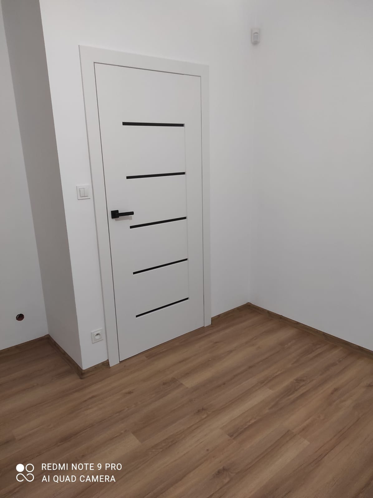 Nowoczesne białe skrzydło drzwiowe z czarnymi, poziomymi intarsjami i czarną klamką, zamontowane w białej futrynie, na tle białych ścian i podłogi z paneli w odcieniach brązu.