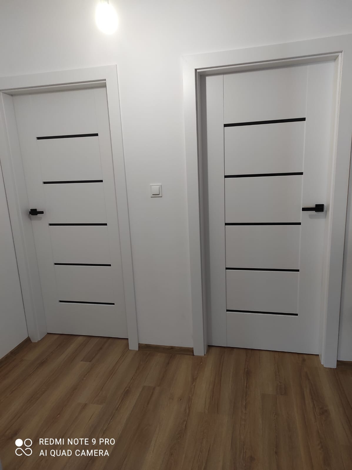 Dwa białe skrzydła drzwiowe z poziomymi, czarnymi wstawkami, zamontowane w białych ościeżnicach, widoczne na tle białej ściany i podłogi imitującej drewno.
