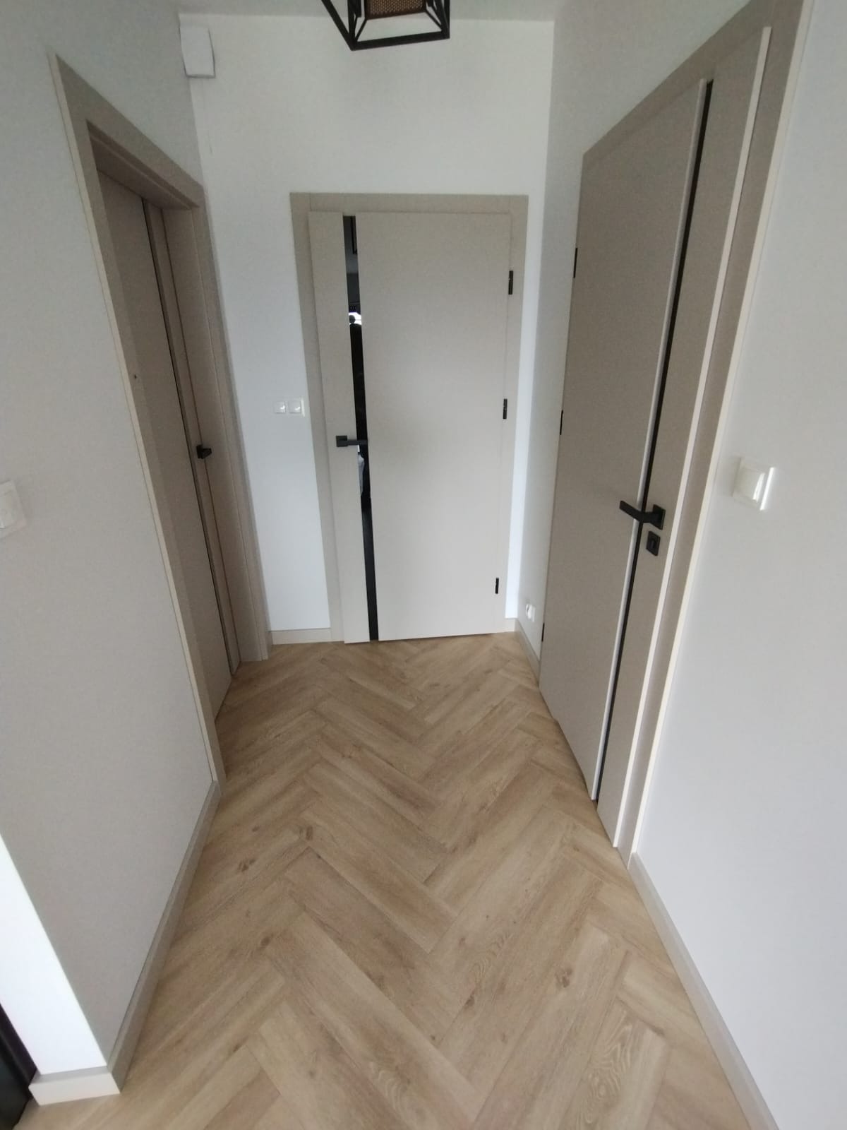 Korytarz z trzema beżowymi drzwiami wewnętrznymi z czarnymi klamkami i panelami ułożonymi w jodełkę.