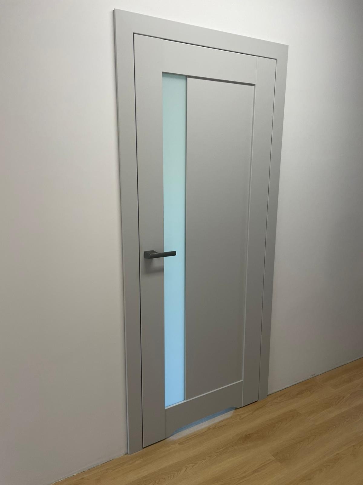 Szara, nowoczesna konstrukcja z prostokątnym przeszkleniem, zamontowana w białej ścianie, widoczna podłoga z paneli.