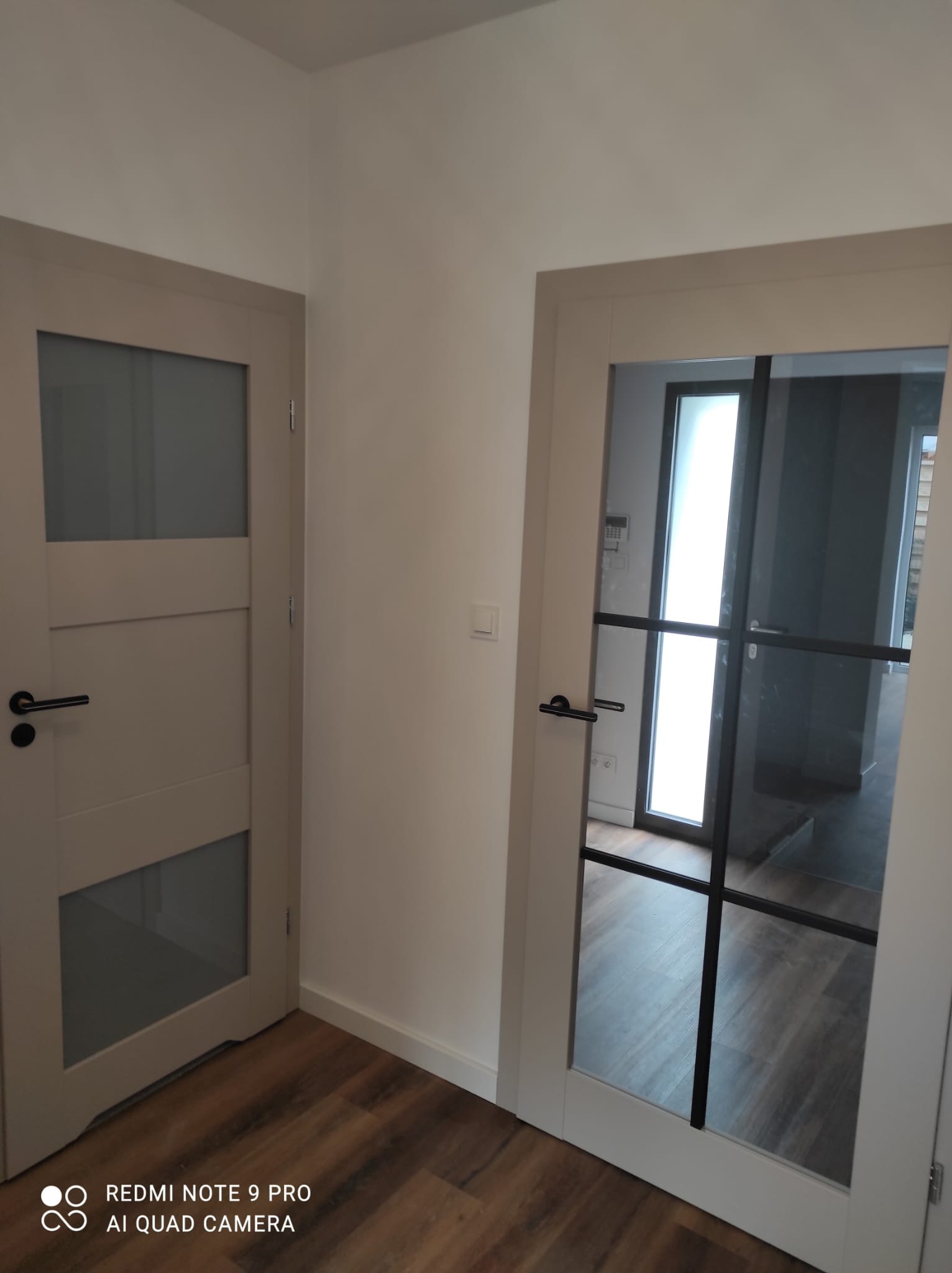 Dwa rodzaje przeszklonych skrzydeł drzwiowych w korytarzu: beżowe z trzema panelami i czarne z podziałami na szklanej tafli.