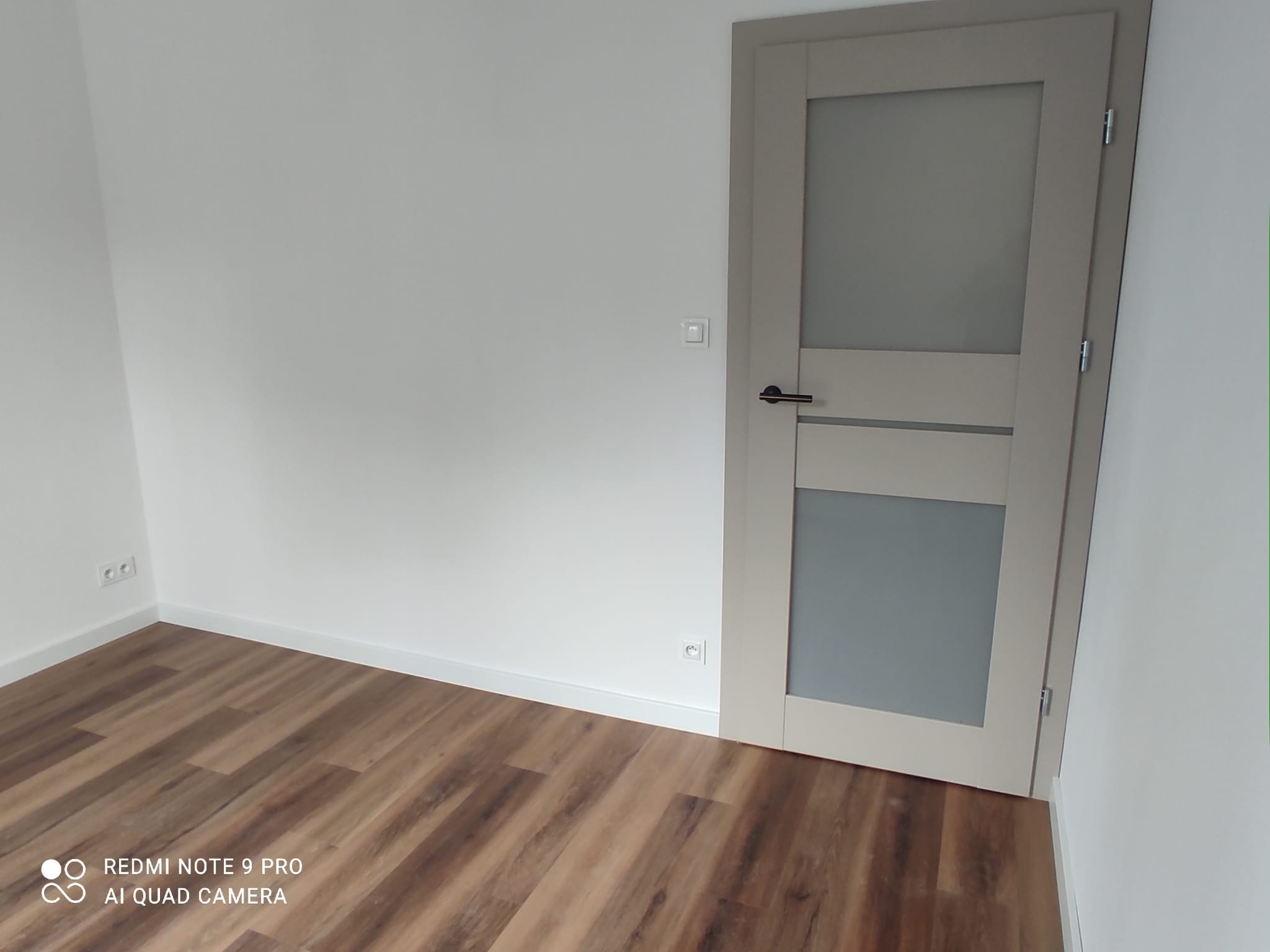 Nowoczesne, beżowe skrzydło drzwiowe z przeszkleniem, zamontowane w białym pomieszczeniu z panelami podłogowymi o wzorze drewna.