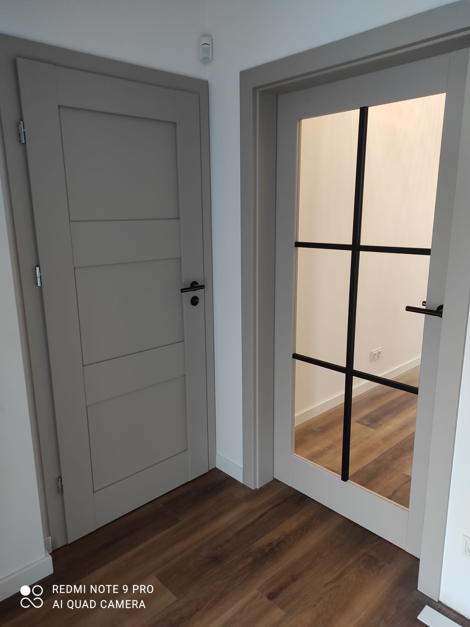 Dwa rodzaje drzwi wewnętrznych: pełne szare drzwi z poziomymi panelami i czarną klamką oraz otwarte drzwi z szybą i czarnymi szprosami, ukazujące podłogę wewnątrz pomieszczenia.