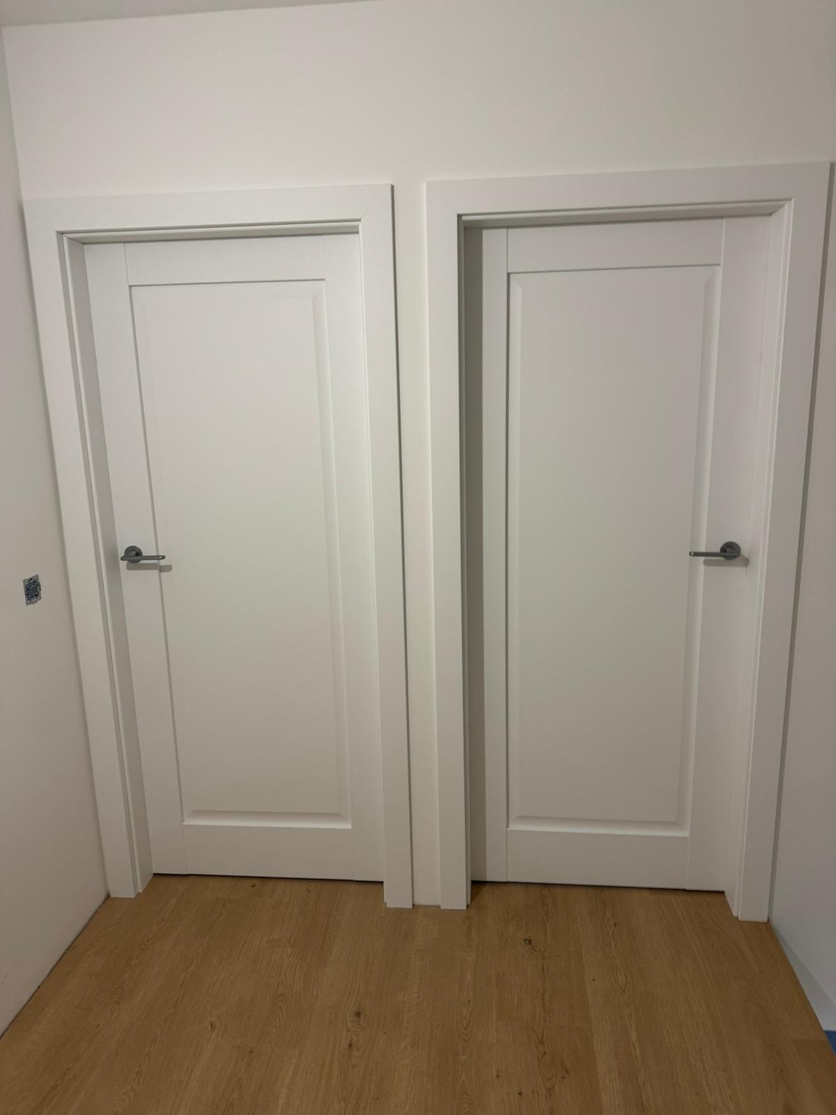 Dwa białe, identyczne skrzydła drzwiowe z panelami i srebrnymi klamkami, zamontowane w białych ościeżnicach, na tle drewnopodobnej podłogi.