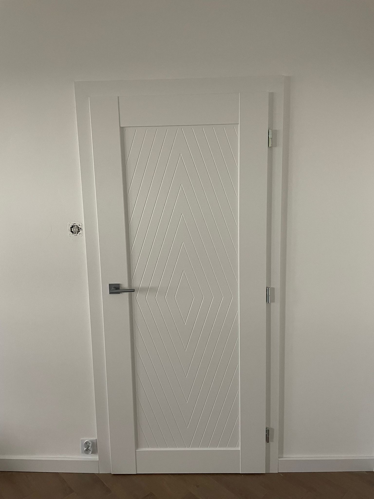 Białe skrzydło z geometrycznym wzorem rombów i srebrną klamką, widoczne gniazdko elektryczne i puszka instalacyjna na ścianie.
