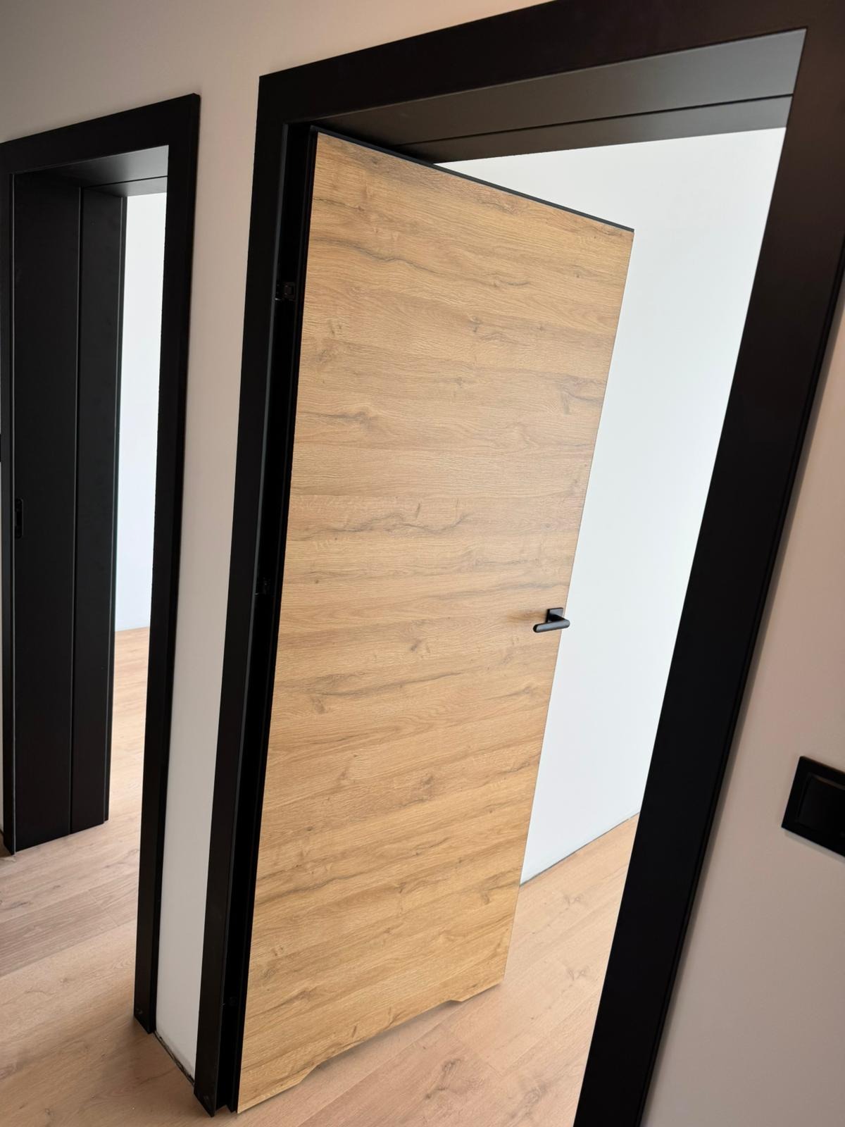 Otwarta, fornirowana skrzydło drzwiowe w czarnej ościeżnicy, widok na sąsiednie pomieszczenie z drugimi drzwiami w czarnej ramie, drewniana podłoga.