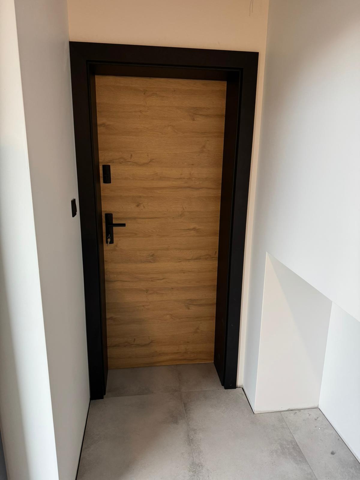 Drewnopodobne skrzydło drzwiowe z czarną ościeżnicą i klamką, widoczne od strony wewnętrznej, z kluczem w zamku.