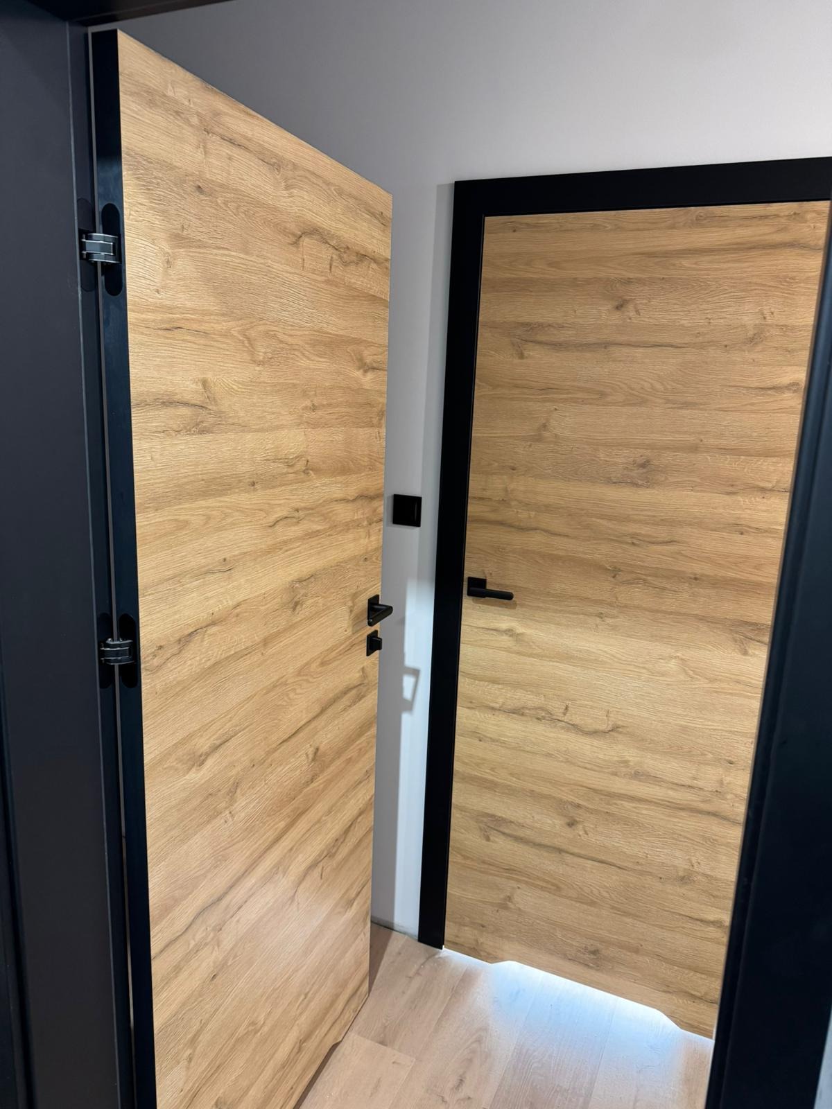 Otwarta dębowa okleina na skrzydle drzwi z czarną ościeżnicą i ukrytymi zawiasami, widoczne drugie skrzydło drzwiowe z czarną klamką i ościeżnicą.