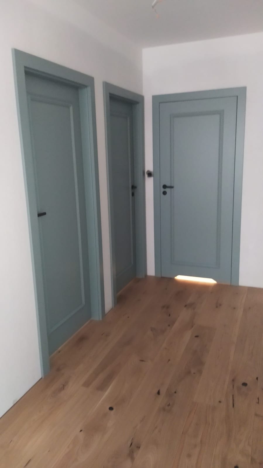 Trzy szare drzwi z czarnymi klamkami zamontowane w białej ścianie, widoczne na tle drewnianej podłogi, z delikatnym oświetleniem padającym spod jednego z nich.