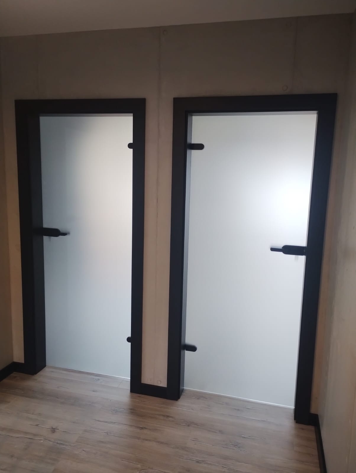 Dwa skrzydła drzwiowe z matowego szkła, osadzone w czarnych ramach, z minimalistycznymi, poziomymi klamkami, umieszczone w jasnym pomieszczeniu z panelami podłogowymi.