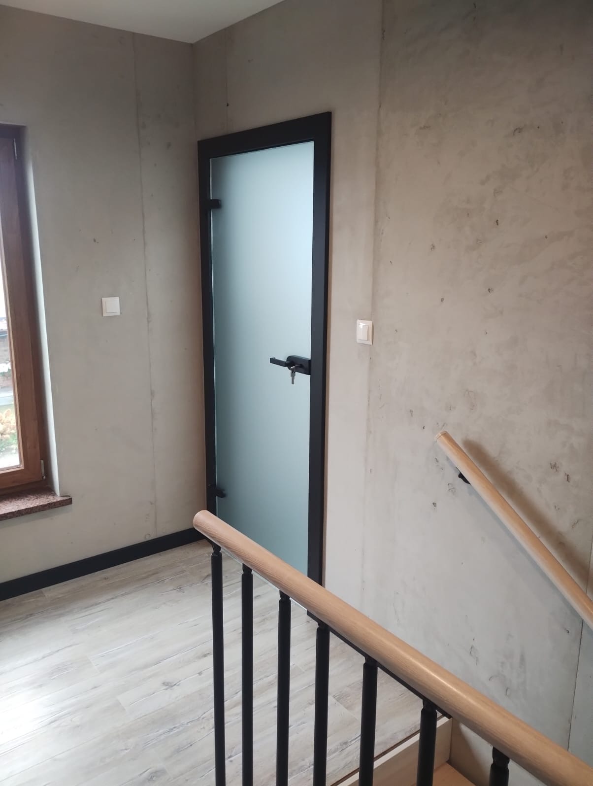 Klatka schodowa z betonowymi ścianami, drewnianą balustradą i drzwiami z matowego szkła w czarnej ramie, widoczne z góry.