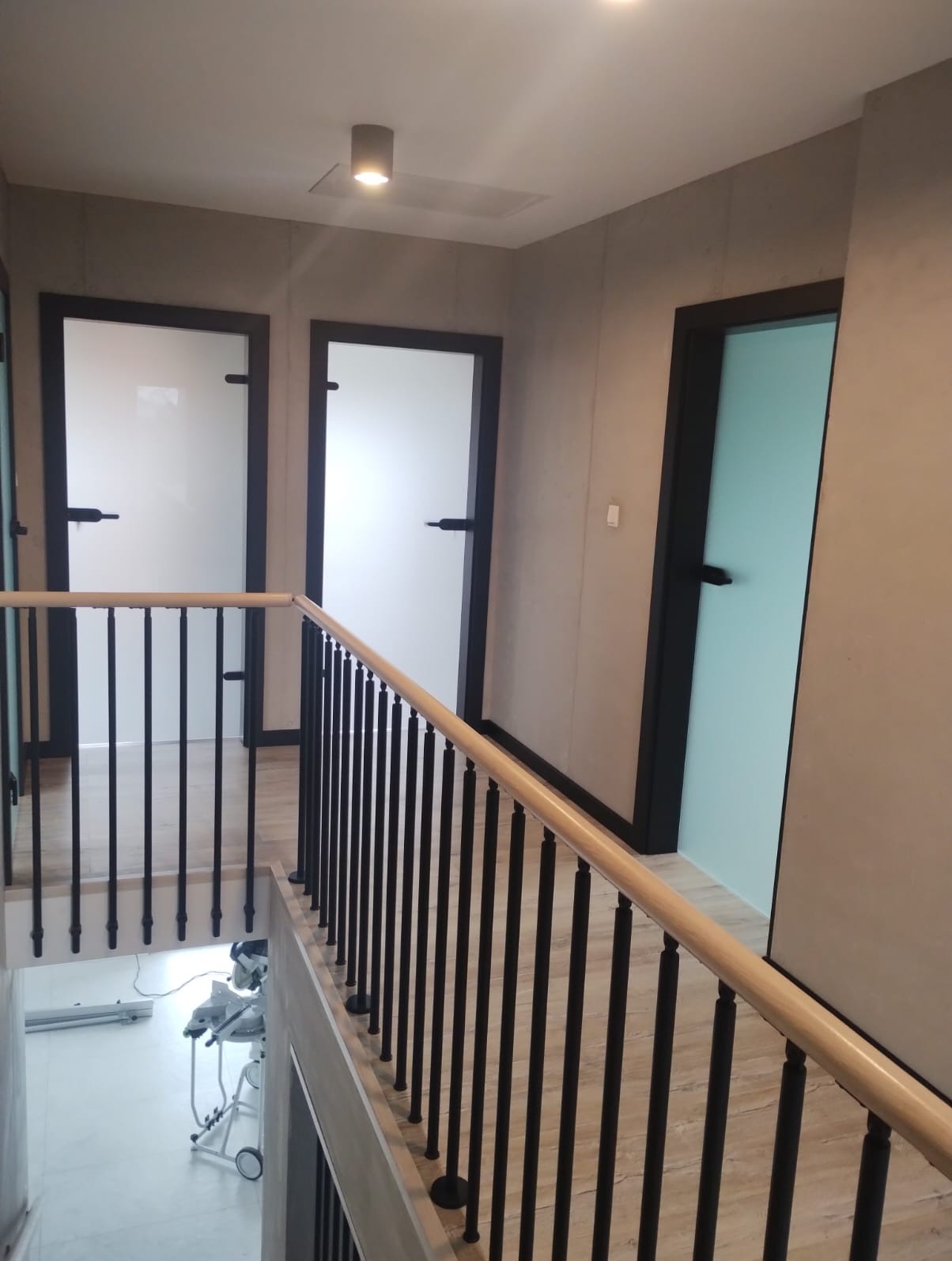 Korytarz z trzema drzwiami o czarnych framugach: dwa białe, jeden turkusowy; balustrada z drewnianą poręczą i czarnymi, pionowymi szczebelkami; na dole widoczna piła ukośnica na stole.