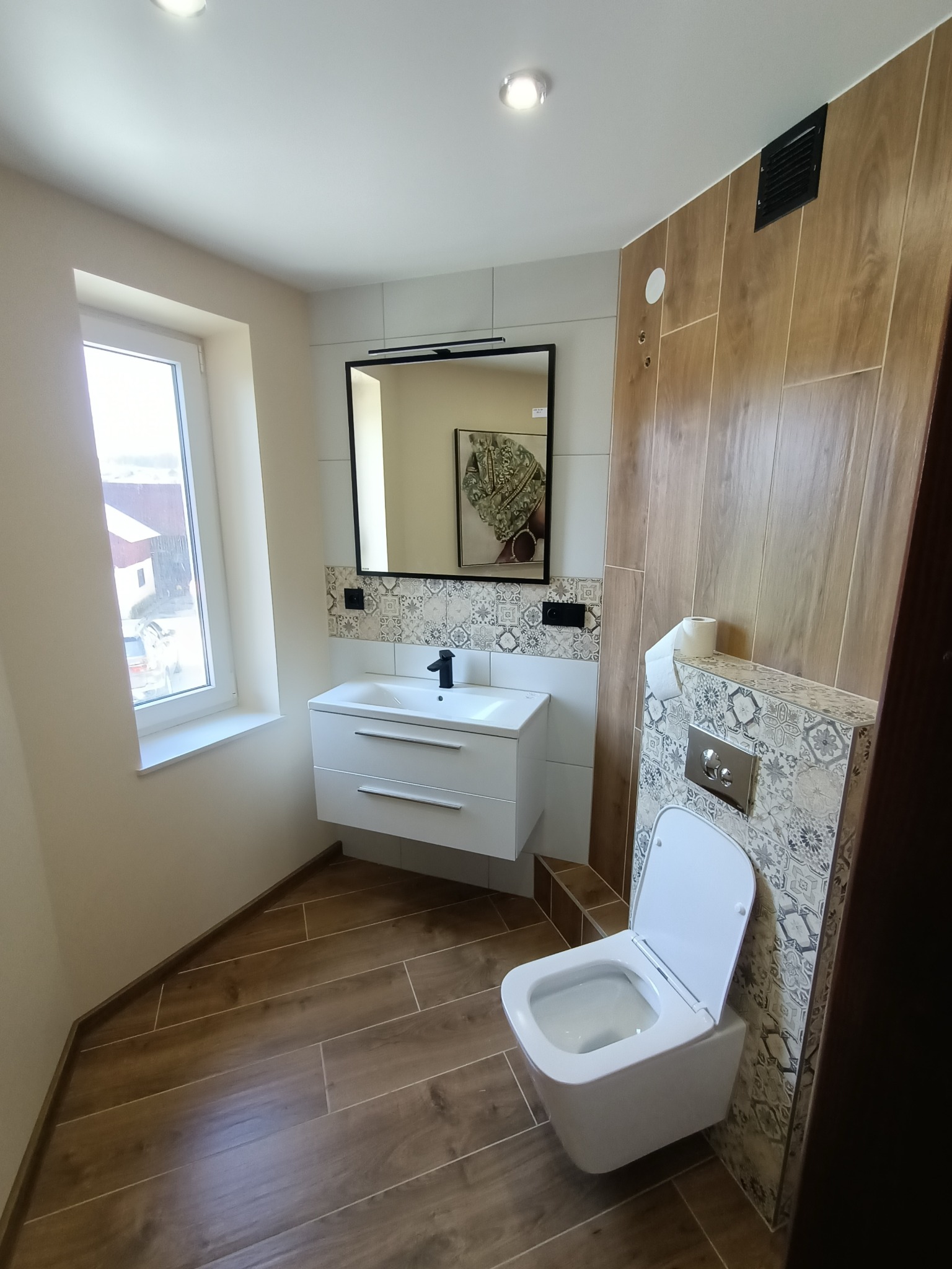 Nowoczesna łazienka z wiszącą toaletą, umywalką z szafką i lustrem w czarnej ramie, wykończona płytkami imitującymi drewno i dekoracyjnymi panelami z motywem marokańskim.