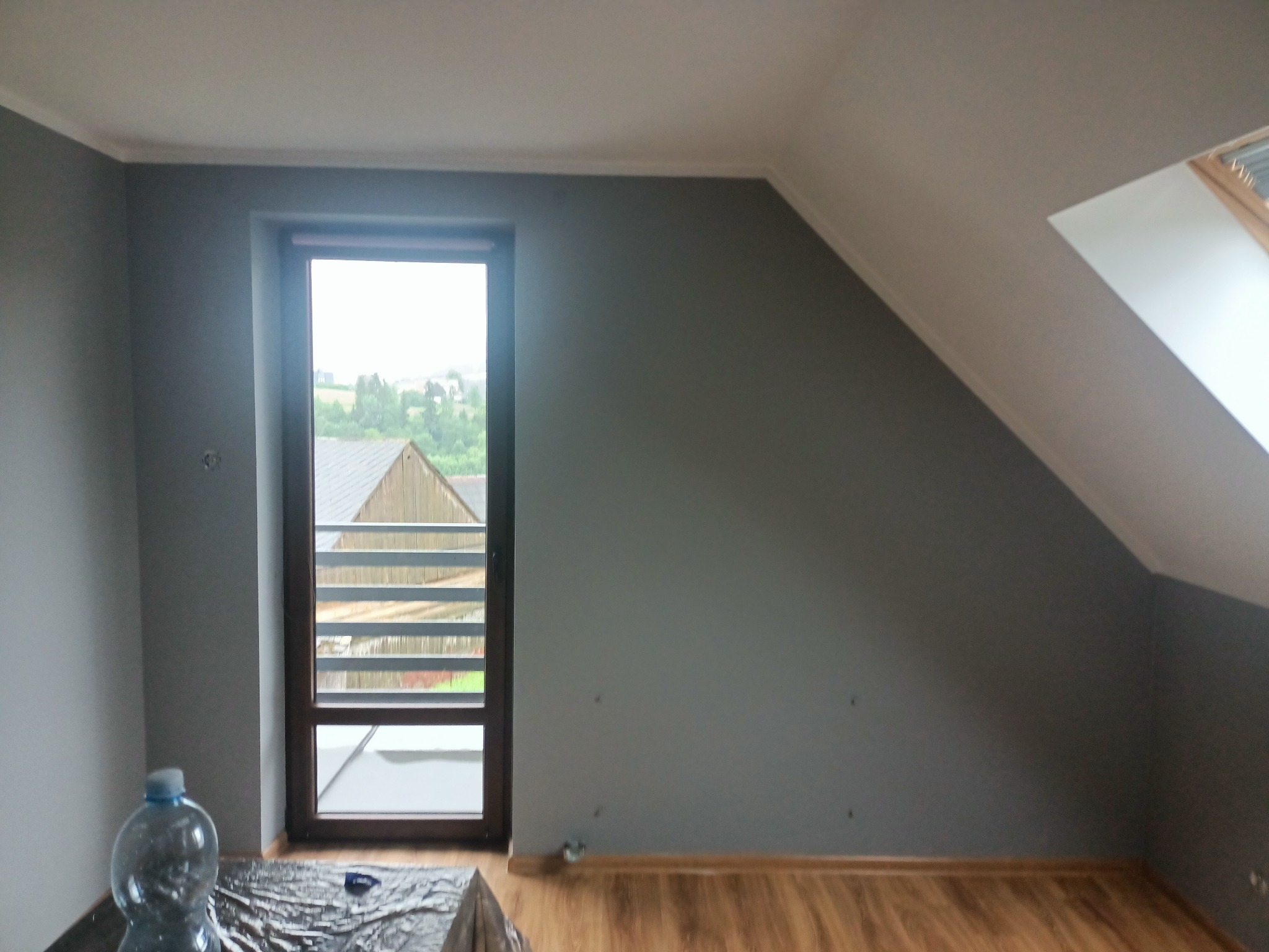Pokój na poddaszu po malowaniu ścian na szaro, widoczne okno dachowe i zwykłe okno z widokiem na zewnątrz, zabezpieczona folią podłoga i butelka wody.