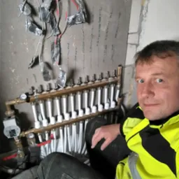 Instalacja rozdzielacza ogrzewania podłogowego z widocznymi przewodami, rurami i manometrami, obok instalator w odblaskowej kurtce.