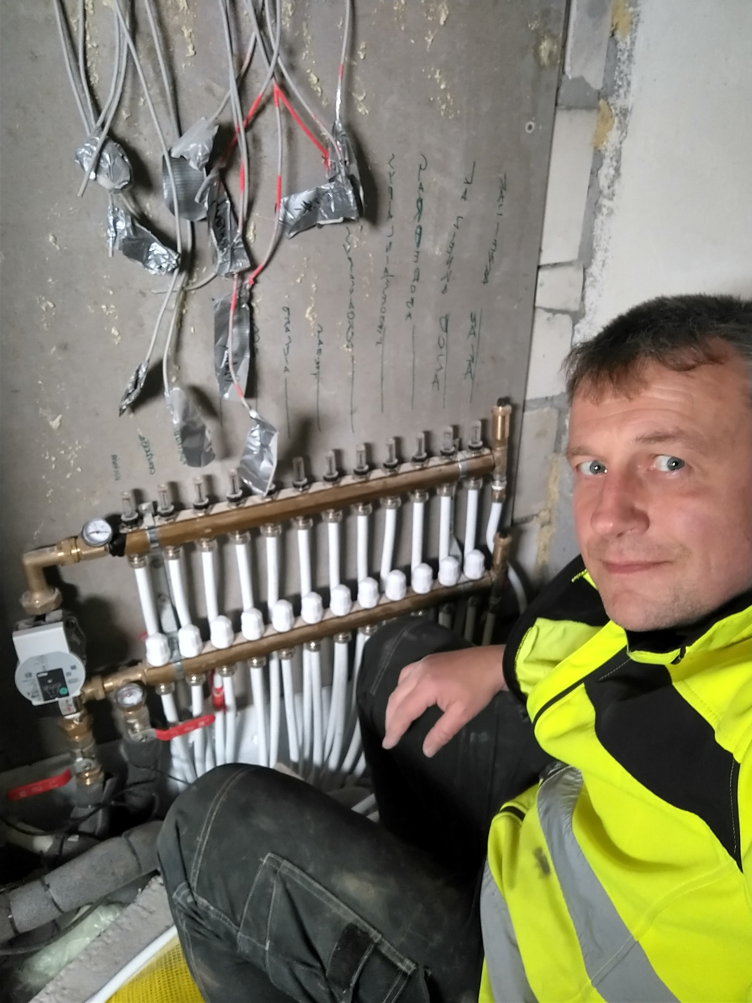 Instalacja rozdzielacza ogrzewania podłogowego z widocznymi przewodami, rurami i manometrami, obok instalator w odblaskowej kurtce.
