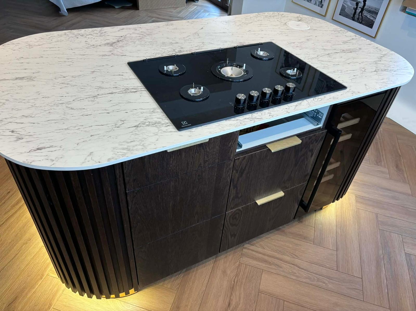 Nowoczesna wyspa kuchenna z marmurowym blatem i wbudowaną płytą grzewczą Electrolux. Ciemne drewno, złote uchwyty, oświetlenie LED u dołu. Parkiet w jodełkę na podłodze.
