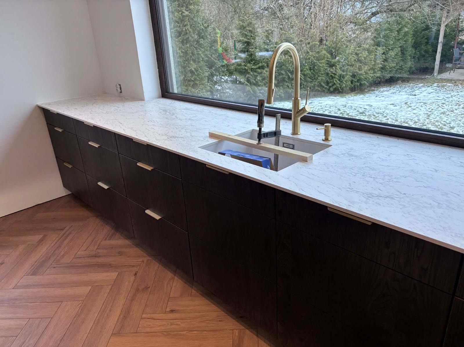 Nowoczesna kuchnia z ciemnymi szafkami, blatem z białego marmuru i złotą armaturą przy oknie z widokiem na ogród. Ułożona podłoga w jodełkę.