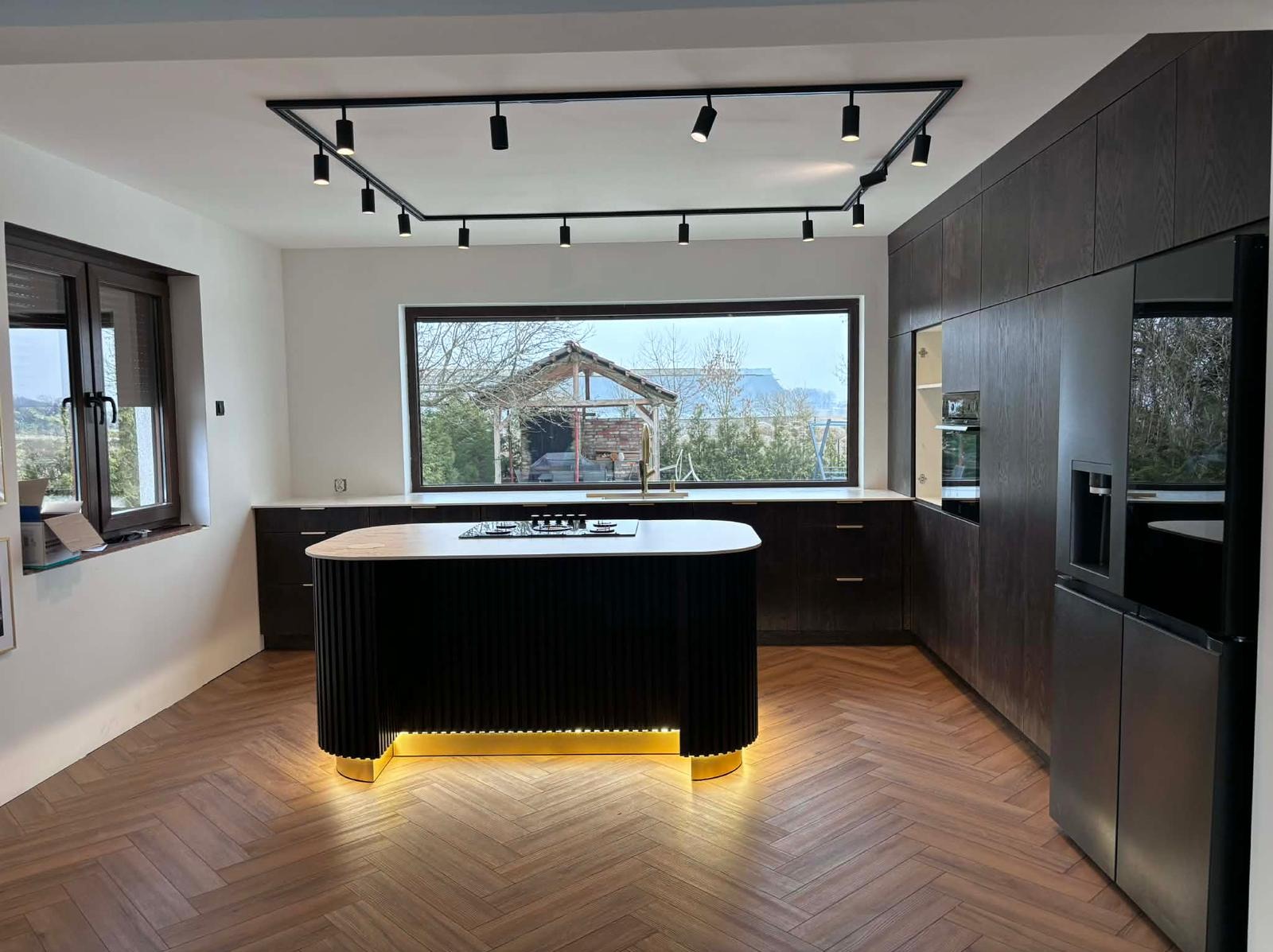 Nowoczesna kuchnia z wyspą oświetloną LED, parkiet w jodełkę, ciemne szafki i duże okno z widokiem na ogród. Oświetlenie punktowe na suficie.