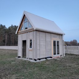 Kemot Tomasz Bienias - Surowy dom szkieletowy w trakcie budowy, z widocznymi panelami ściennymi, oknami i dachem z blachy, otoczony trawnikiem i betonowym ogrodzeniem w tle.