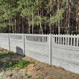 Kemot Tomasz Bienias - Nowo postawione, prefabrykowane ogrodzenie betonowe z dekoracyjnymi przęsłami, imitujące mur z cegły, na tle lasu sosnowego, z widocznymi śladami prac ziemnych i pomarańczowymi kablami na ziemi.