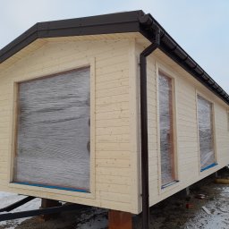Kemot Tomasz Bienias - Nowy, drewniany dom szkieletowy z brązowymi rynnami, okna zabezpieczone folią, ustawiony na bloczkach w zimowej scenerii.