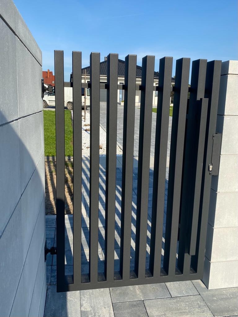 Nowoczesna, grafitowa furtka z pionowych profili, zamontowana między betonowymi słupkami, z widocznym zamkiem i kostką brukową na podjeździe, w tle dom jednorodzinny.