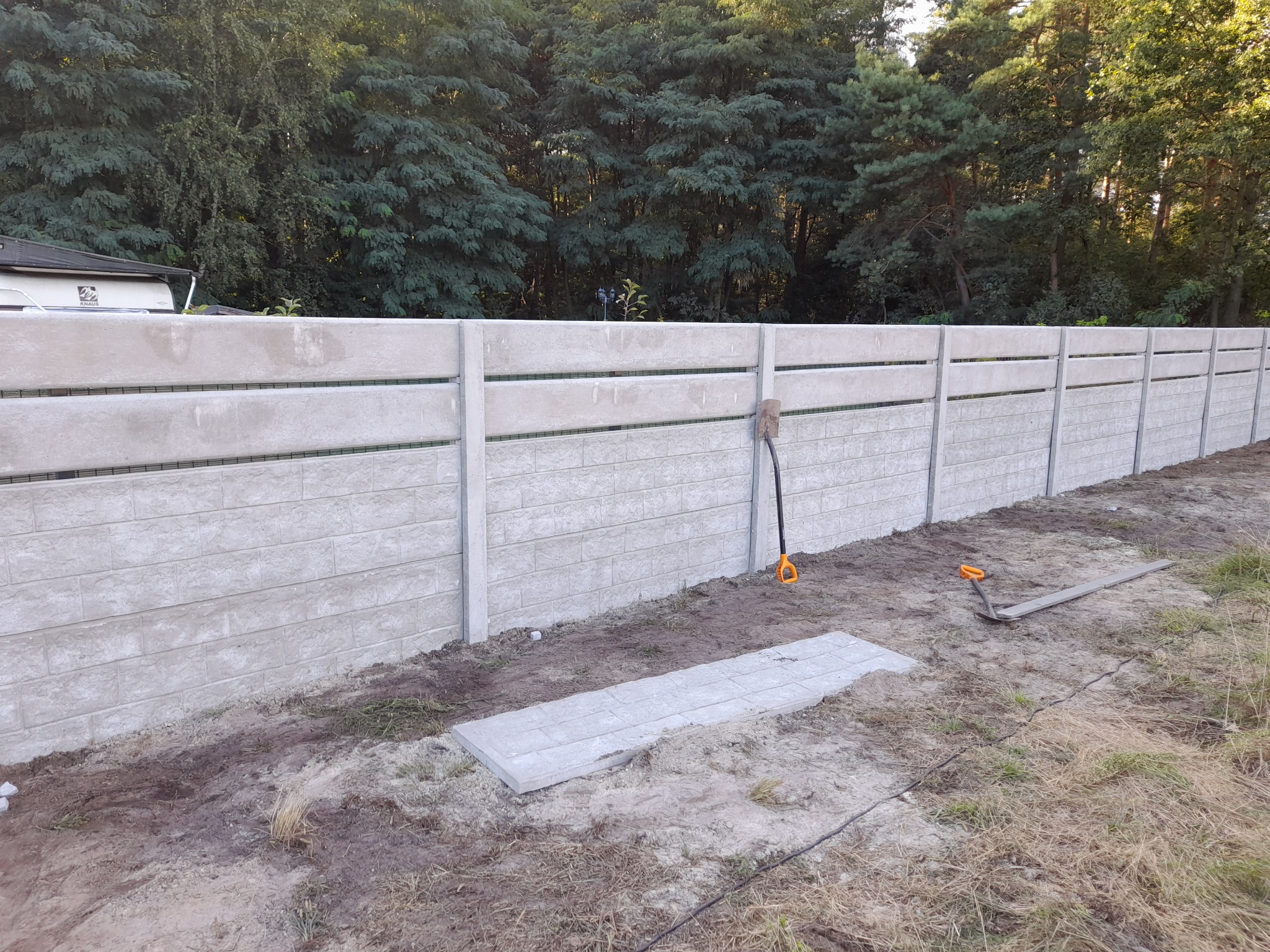 Długie, betonowe ogrodzenie panelowe z fakturą cegły, częściowo ukończone, z widocznymi narzędziami (szpadel i łopata) opartymi o jeden z paneli, na tle drzew i krzewów.