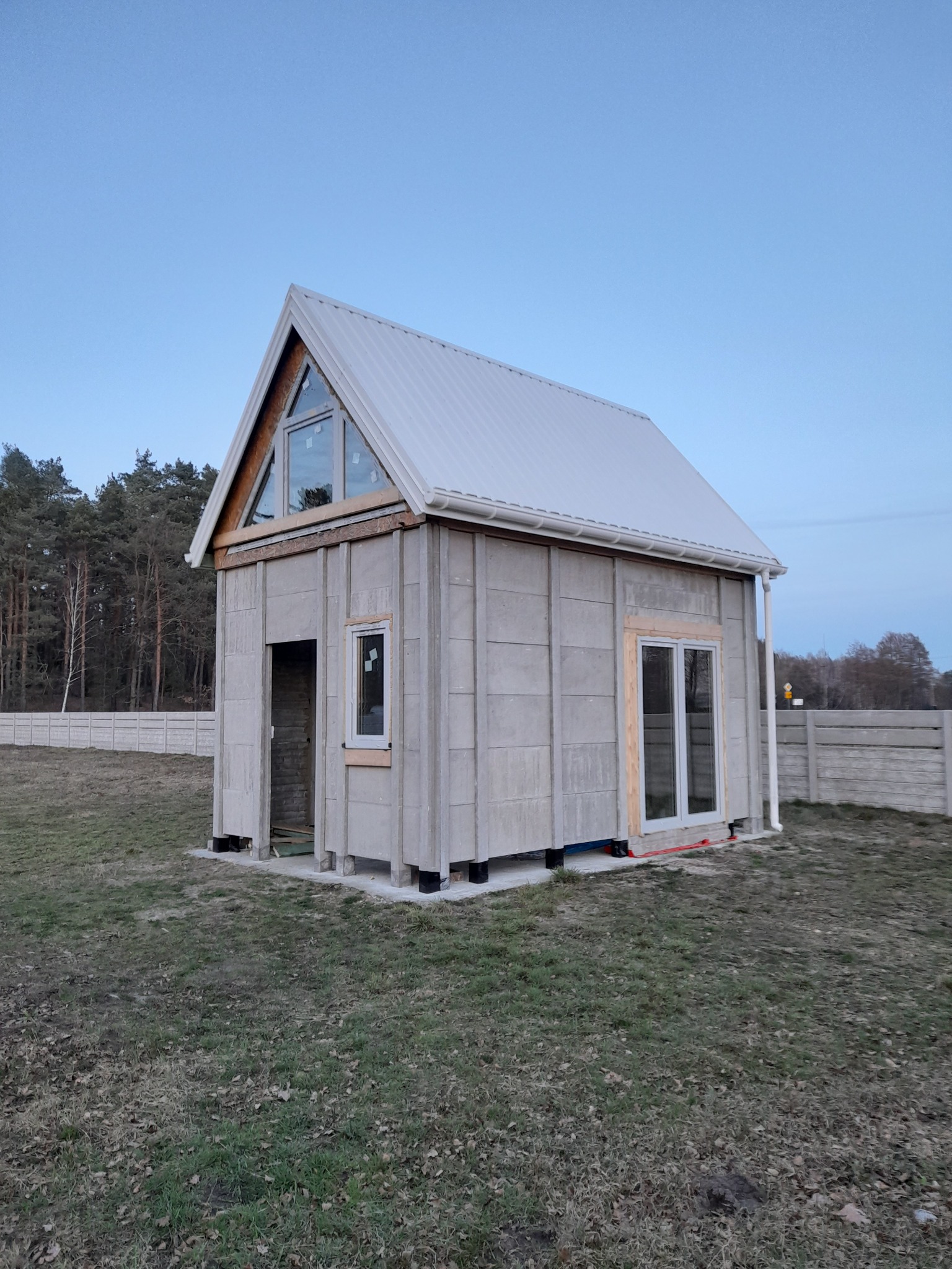 Surowy dom szkieletowy w trakcie budowy, z widocznymi panelami ściennymi, oknami i dachem z blachy, otoczony trawnikiem i betonowym ogrodzeniem w tle.