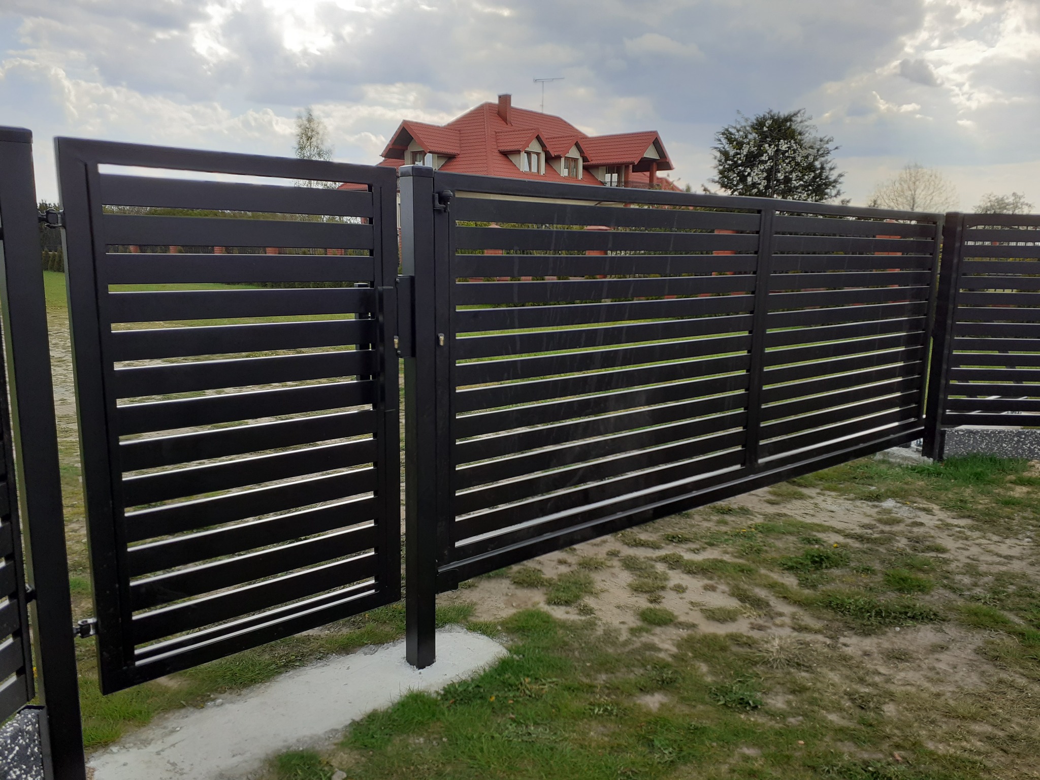 Nowoczesna, pozioma, metalowa brama wjazdowa i ogrodzenie w kolorze antracytowym, z widocznym domem jednorodzinnym z czerwoną dachówką w tle.