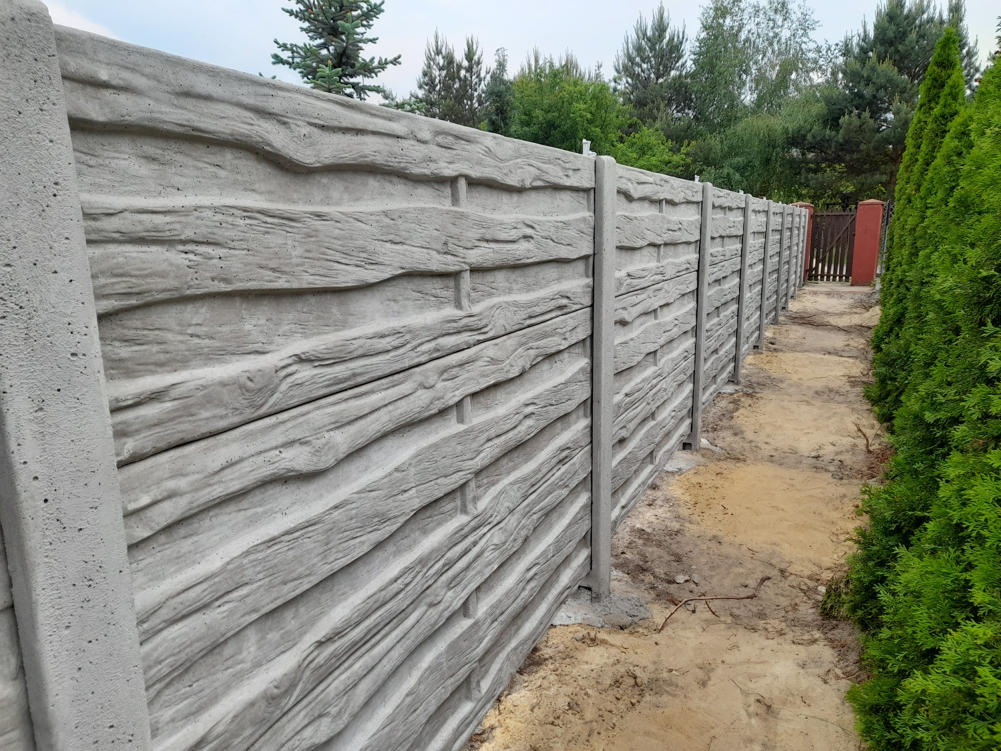 Długi, szary betonowy płot imitujący drewno, ciągnący się wzdłuż piaszczystej ścieżki obok zielonych krzewów i drzew.