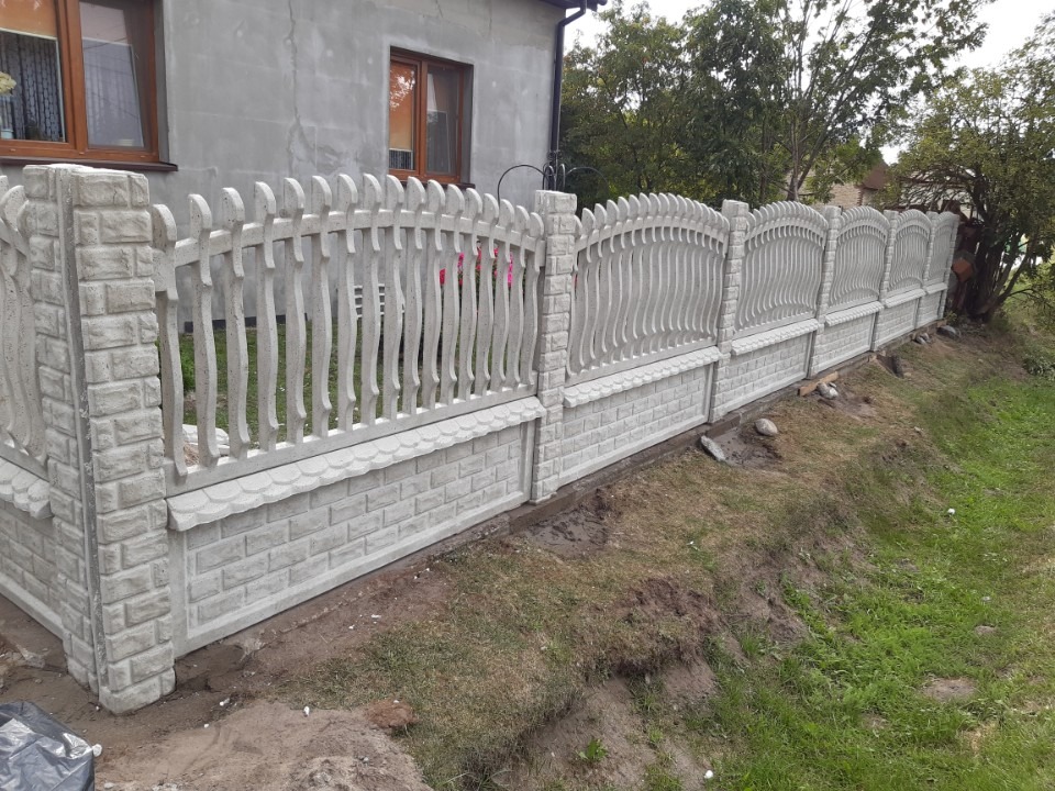 Długi, jasnoszary, betonowy płot z elementami imitującymi cegłę i dekoracyjnymi, ażurowymi panelami, oddzielający posesję z domem od trawnika.