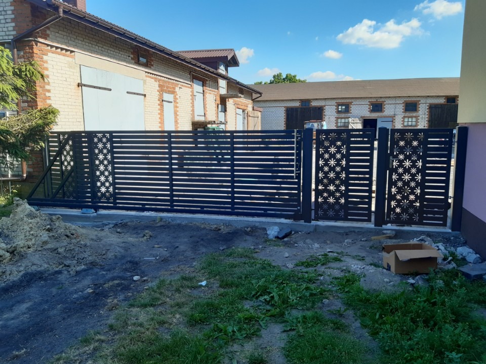 Czarna, przesuwna brama z ażurowym wzorem gwiazd, zamontowana przy posesji z budynkami z cegły, widoczne ślady prac ziemnych.