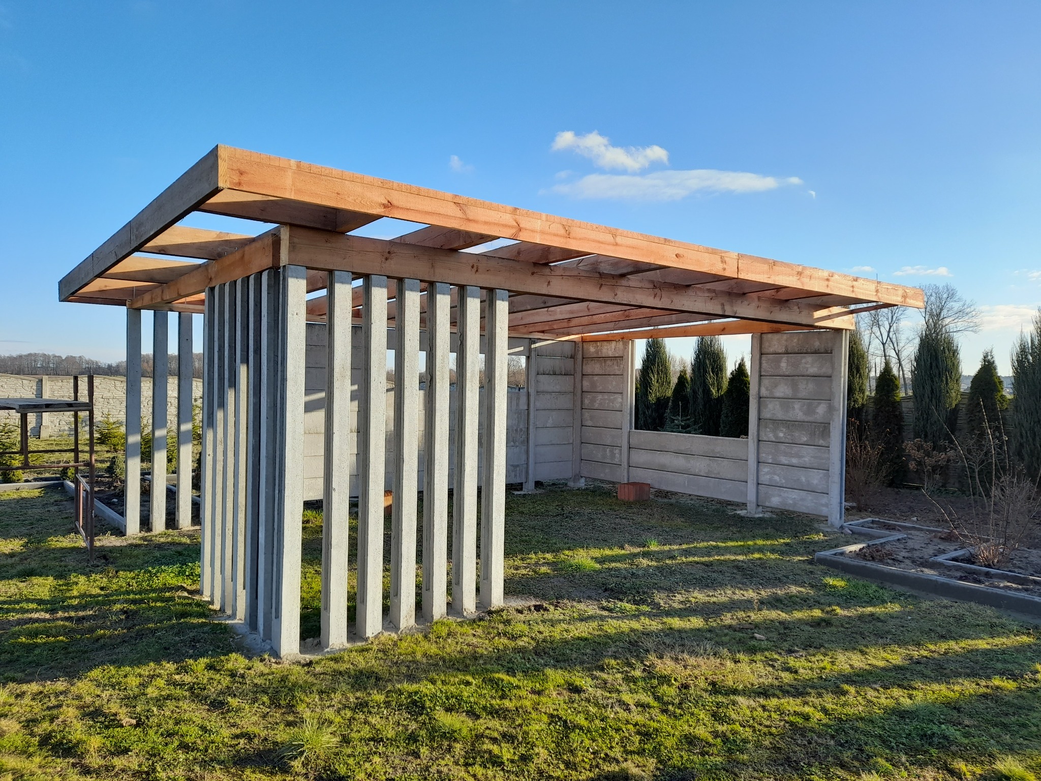 Wznoszona konstrukcja z betonowych słupków i drewnianego szkieletu dachu na trawiastym terenie, w tle drzewa i błękitne niebo.
