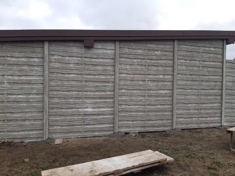 Gotowy fragment ogrodzenia betonowego z brązowym daszkiem, imitujący drewnianą teksturę, świeżo osadzony w ziemi.