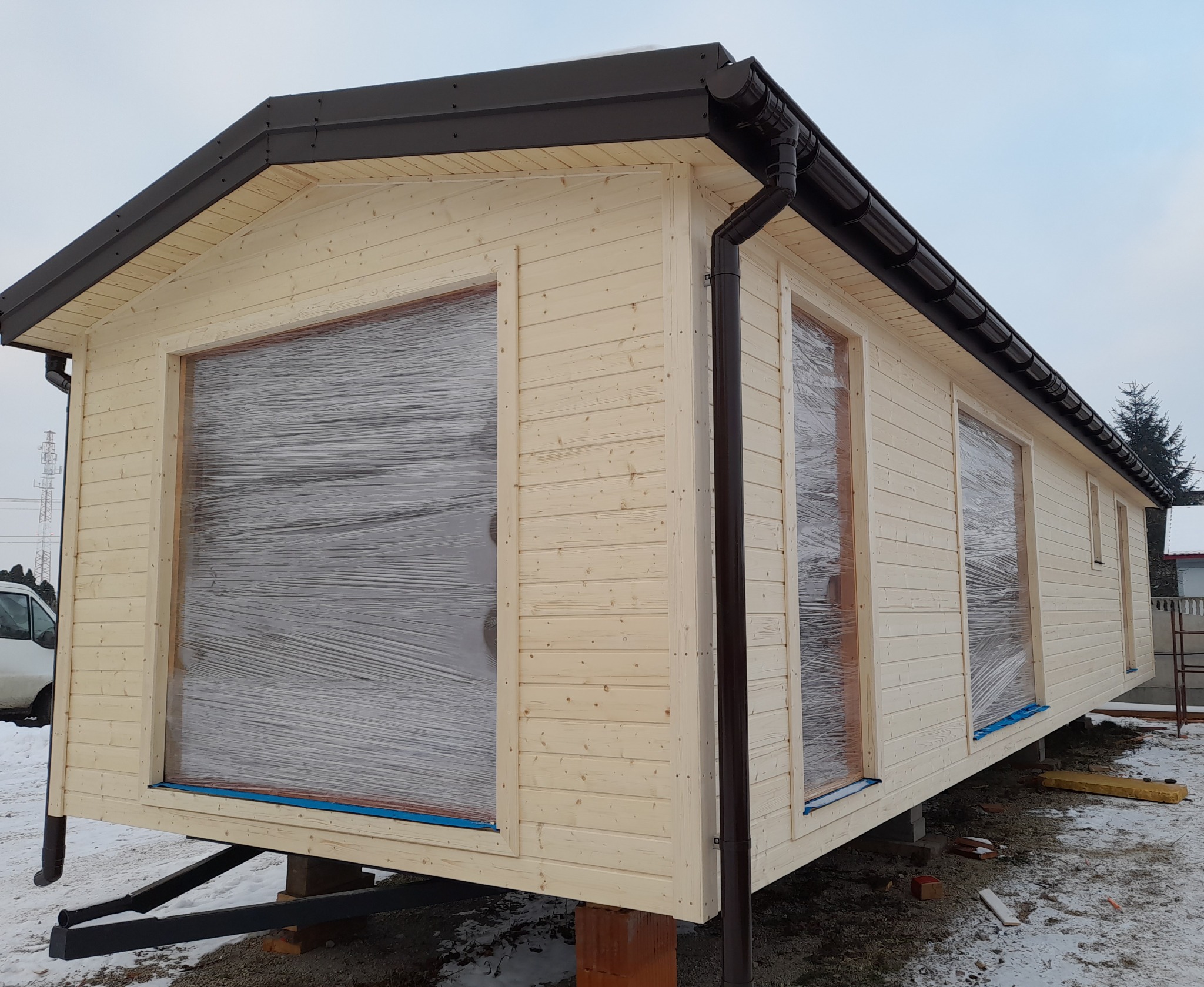 Nowy, drewniany dom szkieletowy z brązowymi rynnami, okna zabezpieczone folią, ustawiony na bloczkach w zimowej scenerii.