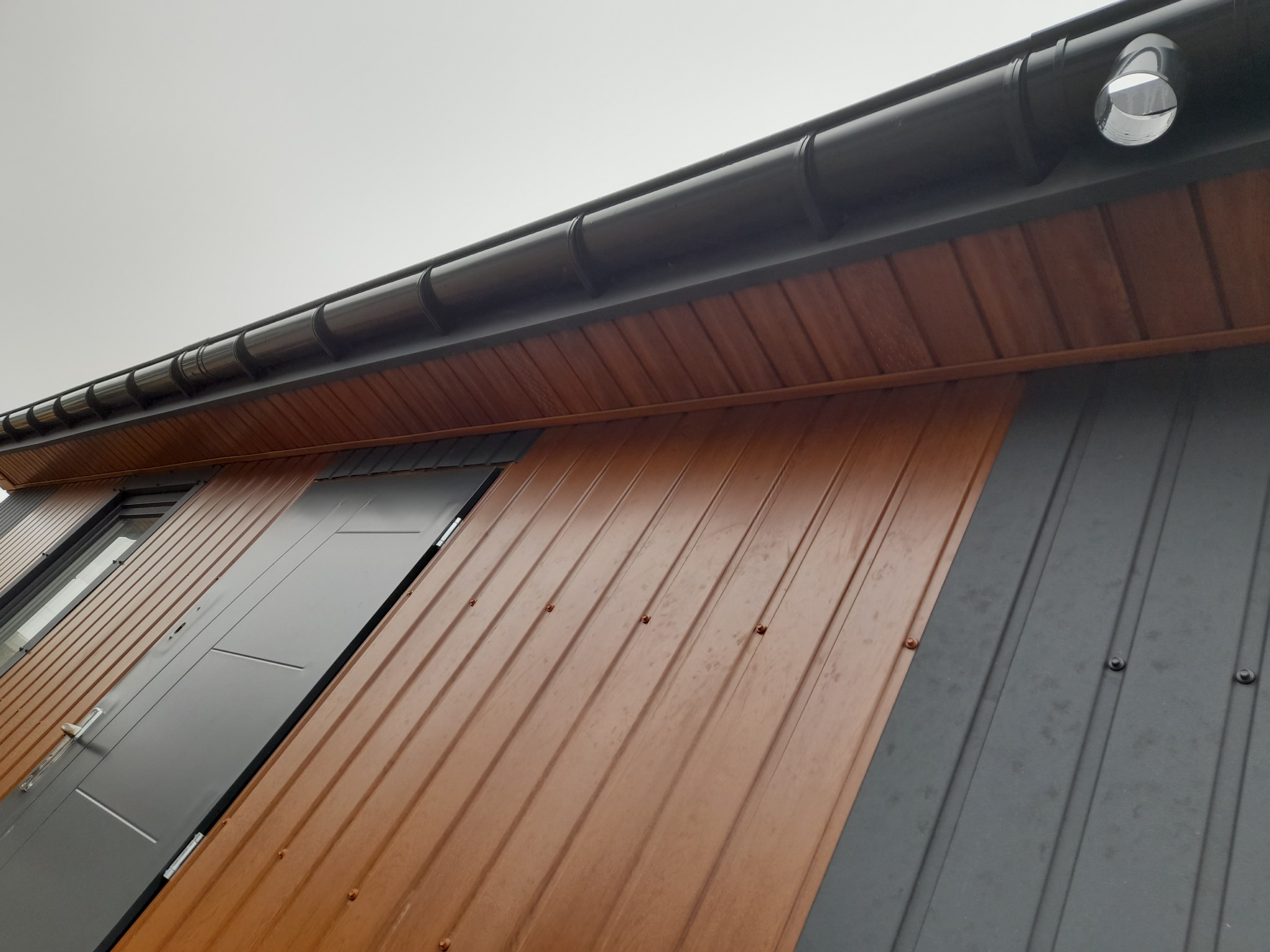 Fragment elewacji budynku z brązową i szarą blachą trapezową, widoczne okno i drzwi wejściowe, orynnowanie z rurą spustową.